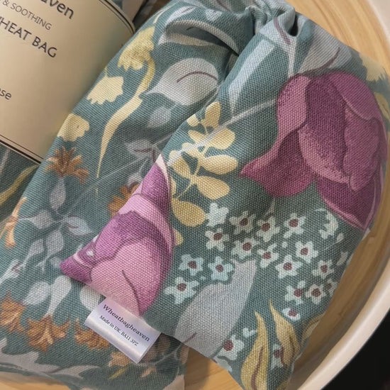 A short video of Two Lavender Wheat bags from WheatBagHeaven with floral design called Cottage Garden in shades of pink, blue and green on a darker sage green base with label that reads WheatBagHeaven, comforting and soothing Lavender Wheat Bag. A natural way to ease those aches and pains also makers tag. The wheat bags are laid on a wooden tray
