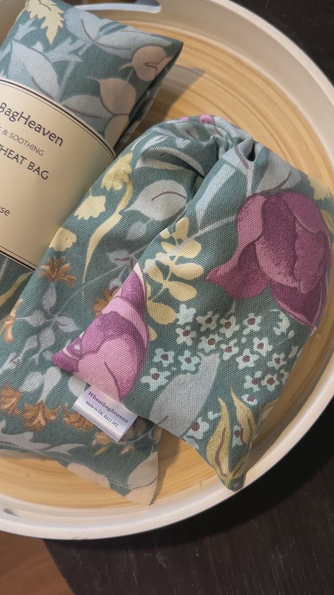 A short video of Two Lavender Wheat bags from WheatBagHeaven with floral design called Cottage Garden in shades of pink, blue and green on a darker sage green base with label that reads WheatBagHeaven, comforting and soothing Lavender Wheat Bag. A natural way to ease those aches and pains also makers tag. The wheat bags are laid on a wooden tray