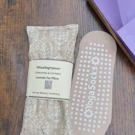 short video of beige yoga socks along side cream and beige eye pillow that as a botanical all over print. The eye pillow has tag and a makers belly band that reads WheatBagHeaven, comforting and soothing Lavender Eye Pillow, calming, relaxing, comforting. The items  are on a wooden table top with lilac packaging in the background.