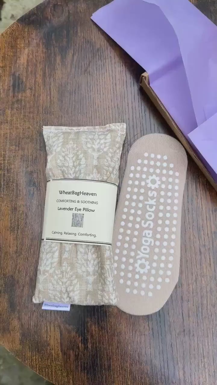 short video of beige yoga socks along side cream and beige eye pillow that as a botanical all over print. The eye pillow has tag and a makers belly band that reads WheatBagHeaven, comforting and soothing Lavender Eye Pillow, calming, relaxing, comforting. The items  are on a wooden table top with lilac packaging in the background.