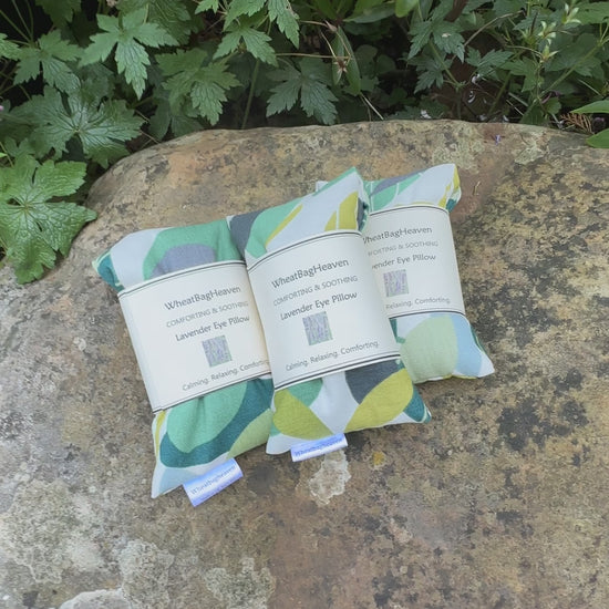 A short video of Three Lavender eye pillows in green spruce cotton fabric, that has an all over leaf pattern in various shades of green on a white base. The eye pillows are laid on a garden stone. They have maker's label that reads WheatBagHeaven comforting and soothing Lavender Eye Pillow, calming. relaxing, comforting
