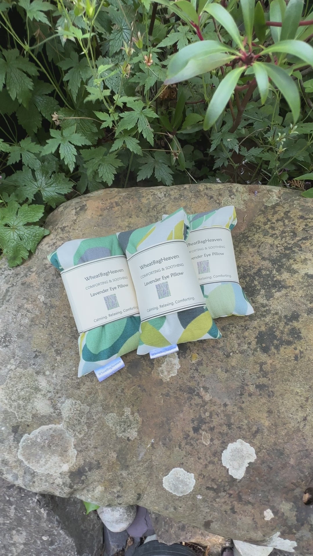 A short video of Three Lavender eye pillows in green spruce cotton fabric, that has an all over leaf pattern in various shades of green on a white base. The eye pillows are laid on a garden stone. They have maker's label that reads WheatBagHeaven comforting and soothing Lavender Eye Pillow, calming. relaxing, comforting