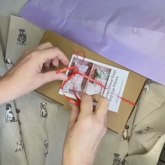 A short video clip of the unpacking of a wheat bag gift. Folded wheat bag washable cover with an all over dog print, Terrier, Boxer, Labrador, Spaniel, Scottie. In shades of grey, brown and cream on a beige base. A plain wheat bag is inside cover. The cover has maker's tag WheatBagHeaven and is laid in a box with lilac tissue paper and ribbon.