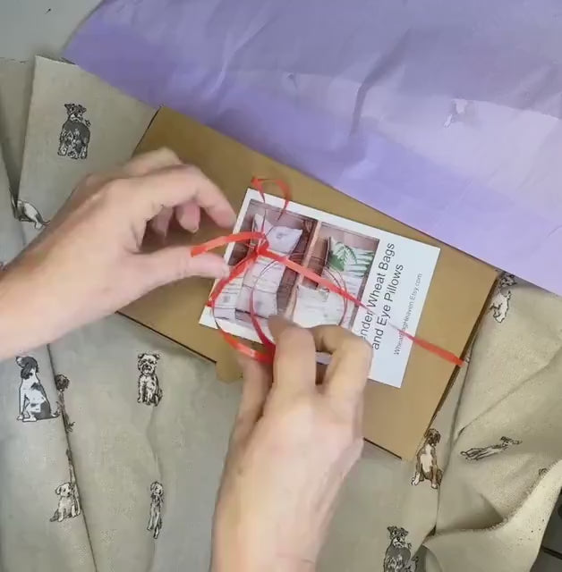 A short video clip of the unpacking of a wheat bag gift. Folded wheat bag washable cover with an all over dog print, Terrier, Boxer, Labrador, Spaniel, Scottie. In shades of grey, brown and cream on a beige base. A plain wheat bag is inside cover. The cover has maker's tag WheatBagHeaven and is laid in a box with lilac tissue paper and ribbon.