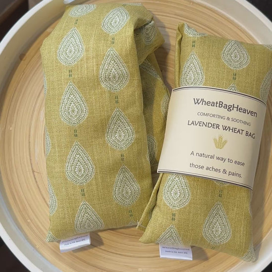 short video of two  lavender wheat bags on a wooden tray. The wheat bag has a paisley teardrop pattern in shades of green and cream with a base colour of ochre. Its labelled "WheatBagHeaven" comforting and soothing lavender wheat bag. A natural way to ease those aches and pains. Also a tag that reads WheatBagHeaven