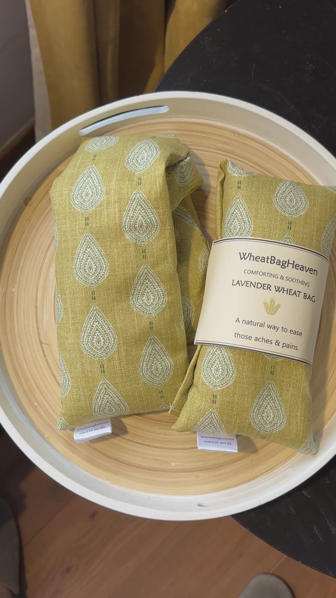 short video of two  lavender wheat bags on a wooden tray. The wheat bag has a paisley teardrop pattern in shades of green and cream with a base colour of ochre. Its labelled "WheatBagHeaven" comforting and soothing lavender wheat bag. A natural way to ease those aches and pains. Also a tag that reads WheatBagHeaven