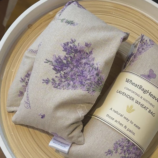 a short video of Two Wheat bag with lavender floral bouquet print and label that reads WheatBagHeaven comforting and soothing Lavender Wheat Bag. A natural way to ease those aches and pains. The wheat bags are laid in a wooden tray
