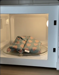 Wheat bag from WheatBagHeaven sat on a turntable in a microwave for heating. The wheat bag has a teal blue base with pink and cream paisley teardrop print and a tag that reads 'WheatBagHeaven'